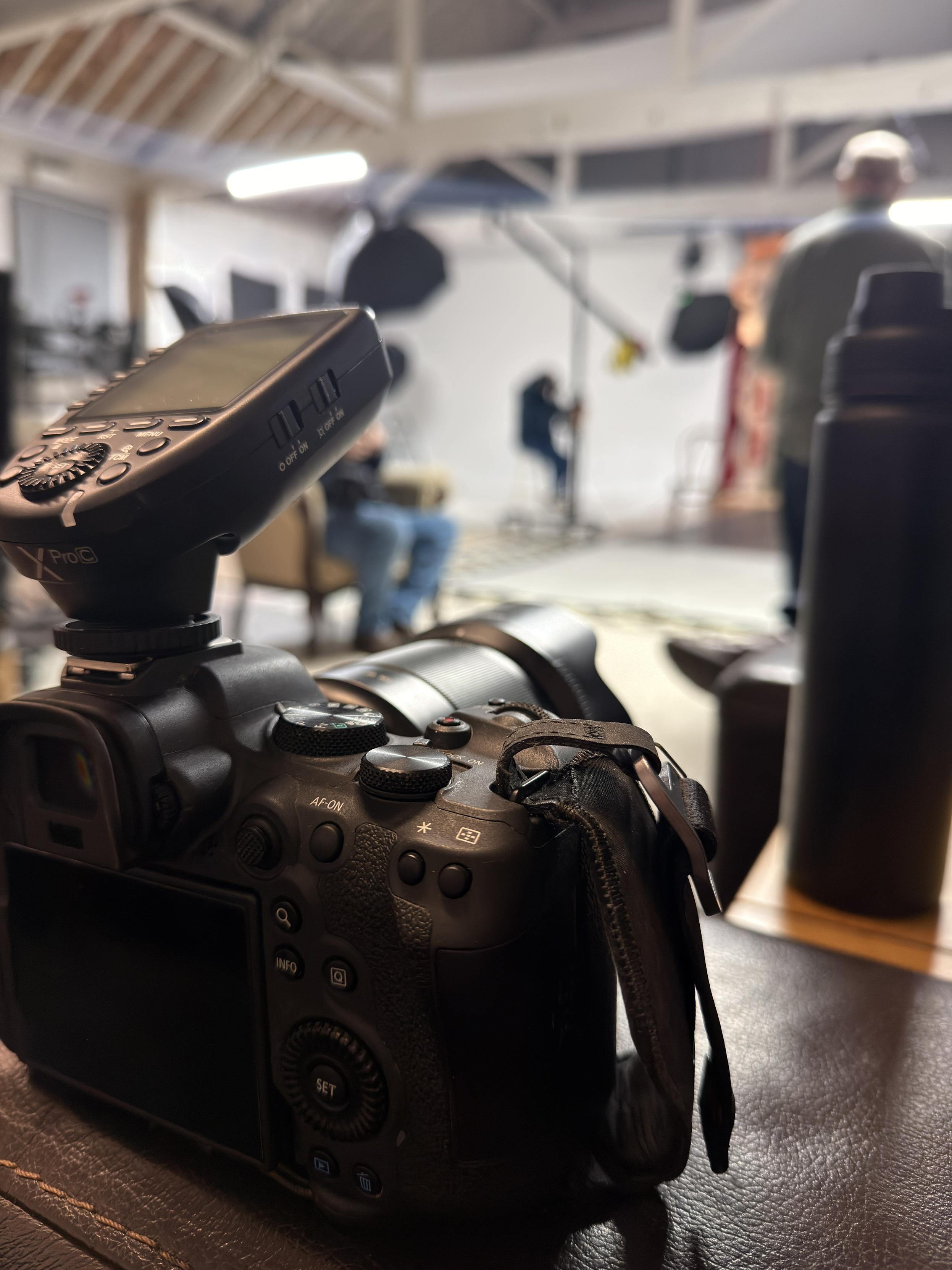 A camera with flash trigger sits on the flat arm of a sofa, in the background out of focus is a photo studio with lights rigged up on the set
