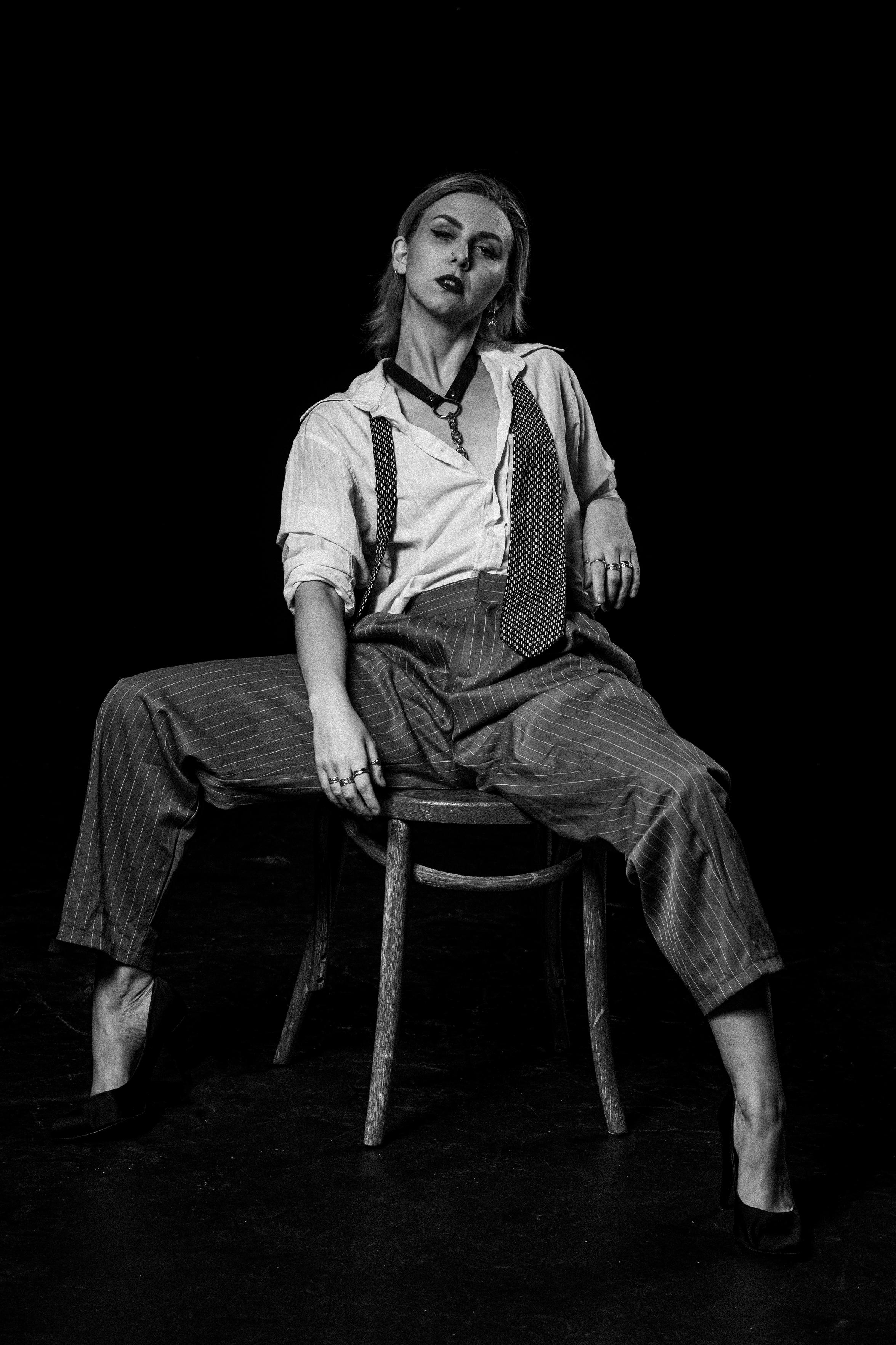 This black and white image captures a person sat on a wooden chair. They're in vintage-style clothes with a loose white shirt, pinstripe trousers, an undone tie draped around their neck, and high-heel shoes. Their pose gives a sense of confidence and an "I don't care what you think" attitude.