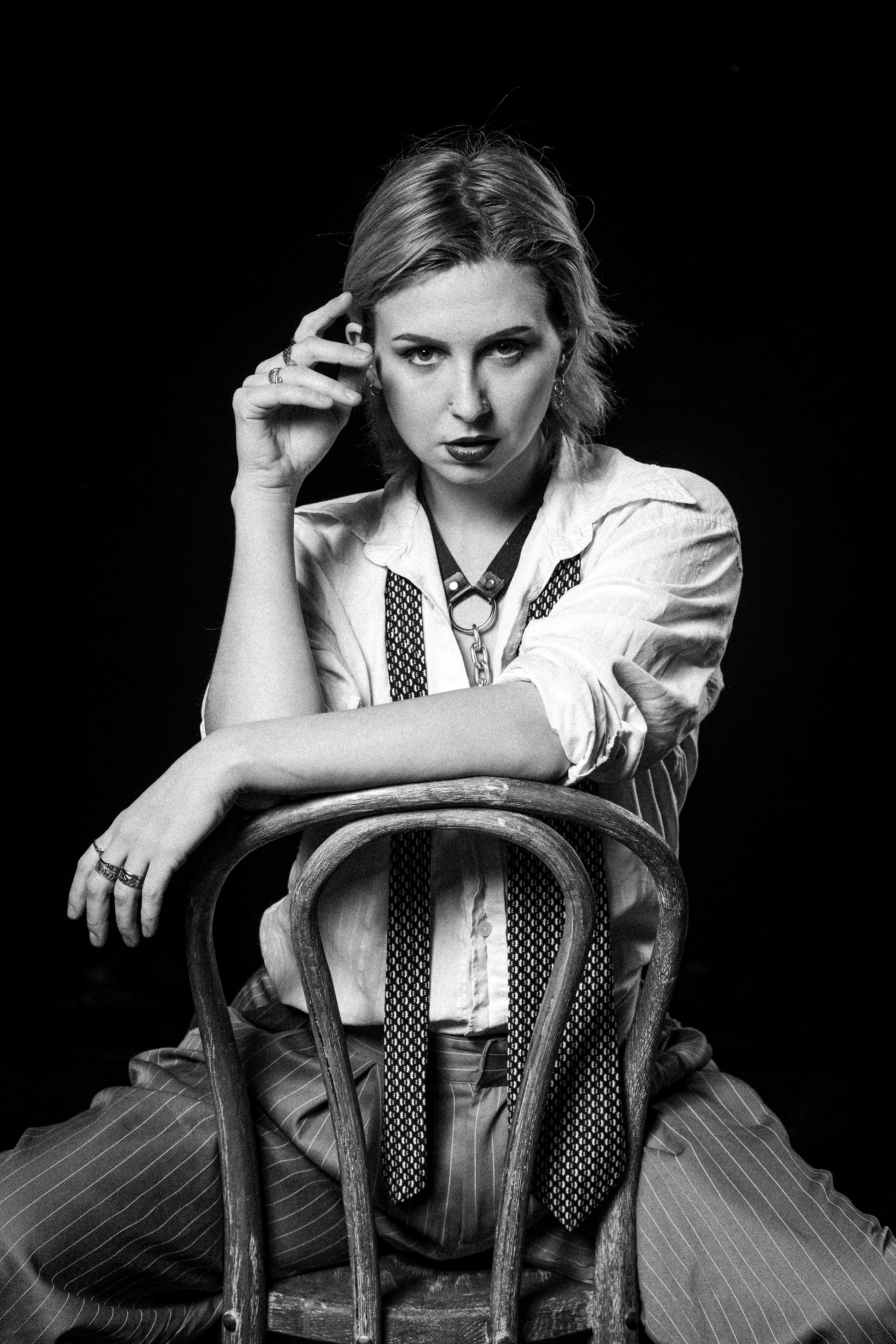 This black and white image captures a person looking directly at the camera, sat on a wooden chair backwards. They're in vintage-style clothes with a loose white shirt, pinstripe trousers, an undone tie draped around their neck. One of their arms reaches up to their face, while the other rests on the back of the chair. It has a sense of confidence and an "I don't care what you think" attitude.
