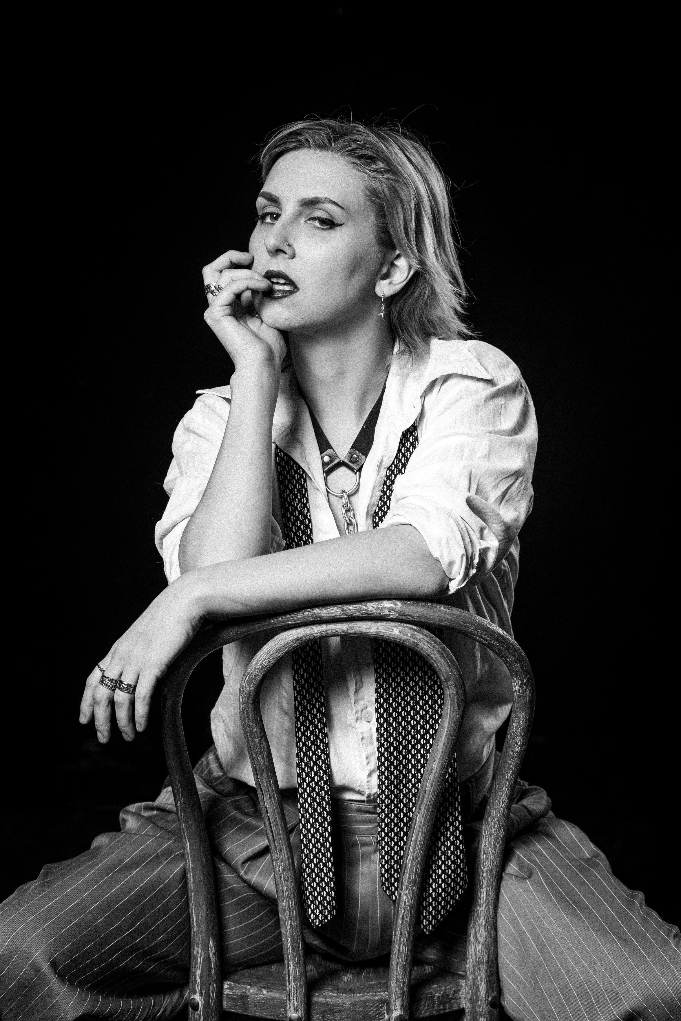This black and white image captures a person looking directly at the camera, sat on a wooden chair backwards. They're in vintage-style clothes with a loose white shirt, pinstripe trousers, an undone tie draped around their neck. One of their arms reaches up to their face with their fingers touching their lips, almost seductively, while the other rests on the back of the chair. It has a sense of confidence and an "I don't care what you think" attitude.