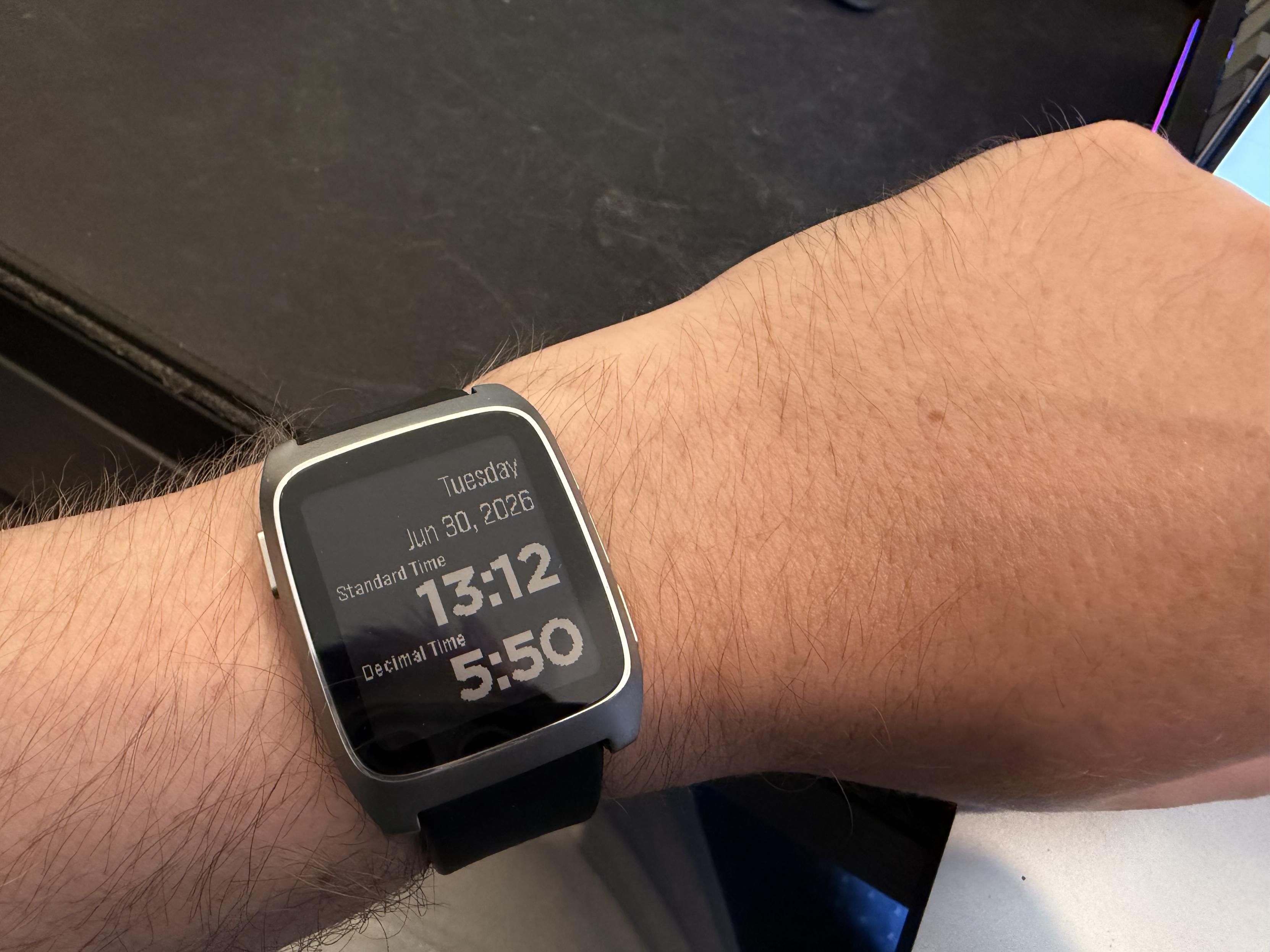 A pebble time 2 watch sits on my wrist, it's a silver case with a black band. The watch face shows a standard time of 13:12 and a decimal time of 5:50